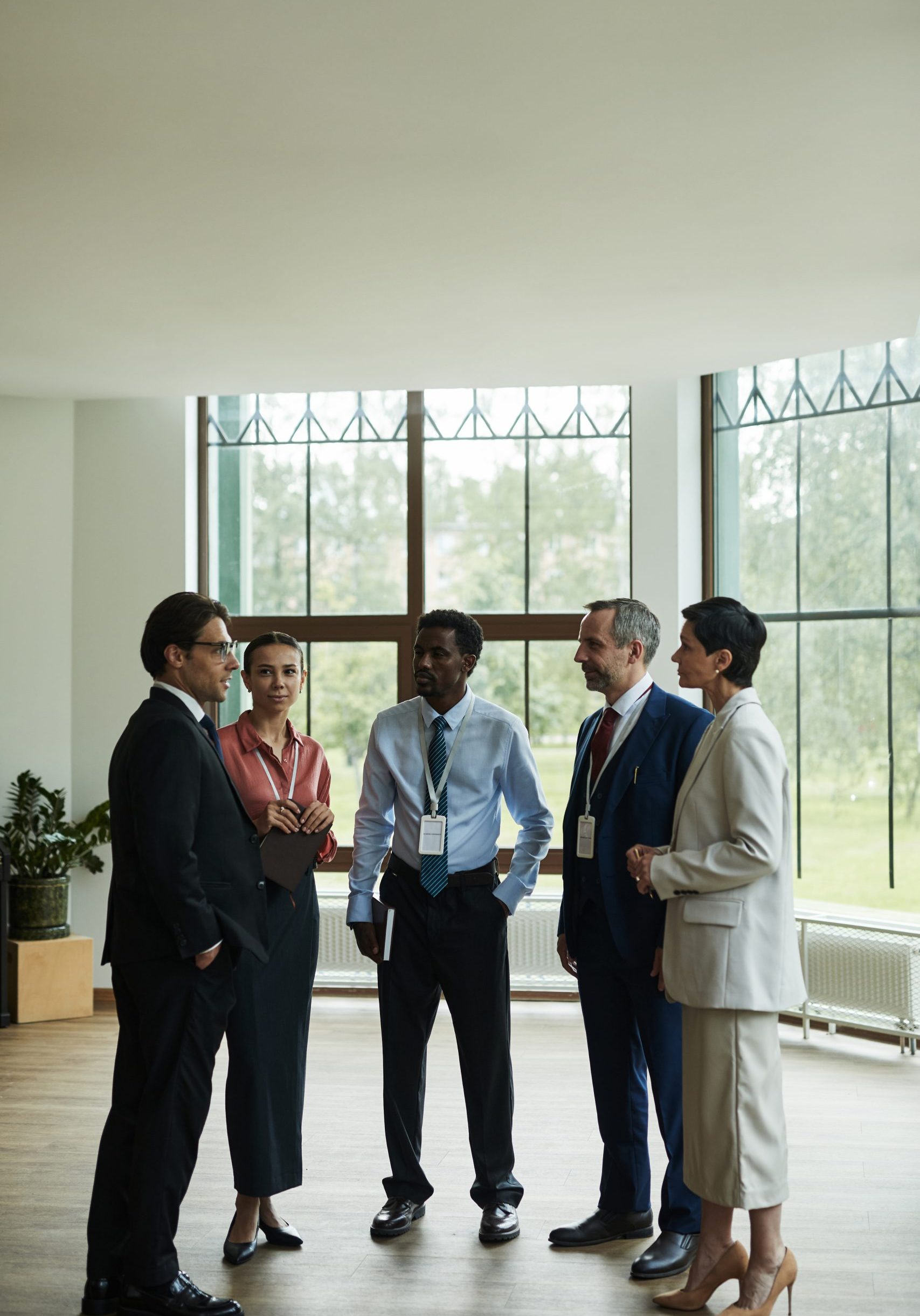 Group of middle aged multiethnic business professionals standing together discussing work in modern office space, all wearing conference badges and holding notebooks or folders